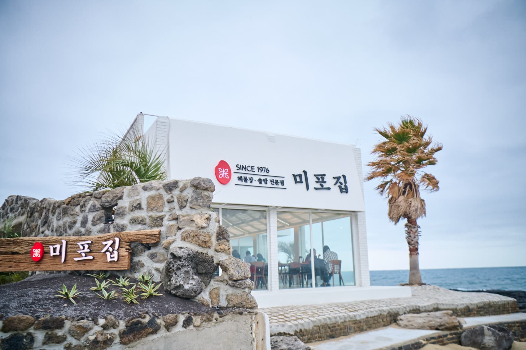 현무암 담장 위 미포집 간판과 야자수, 그 옆의 바다