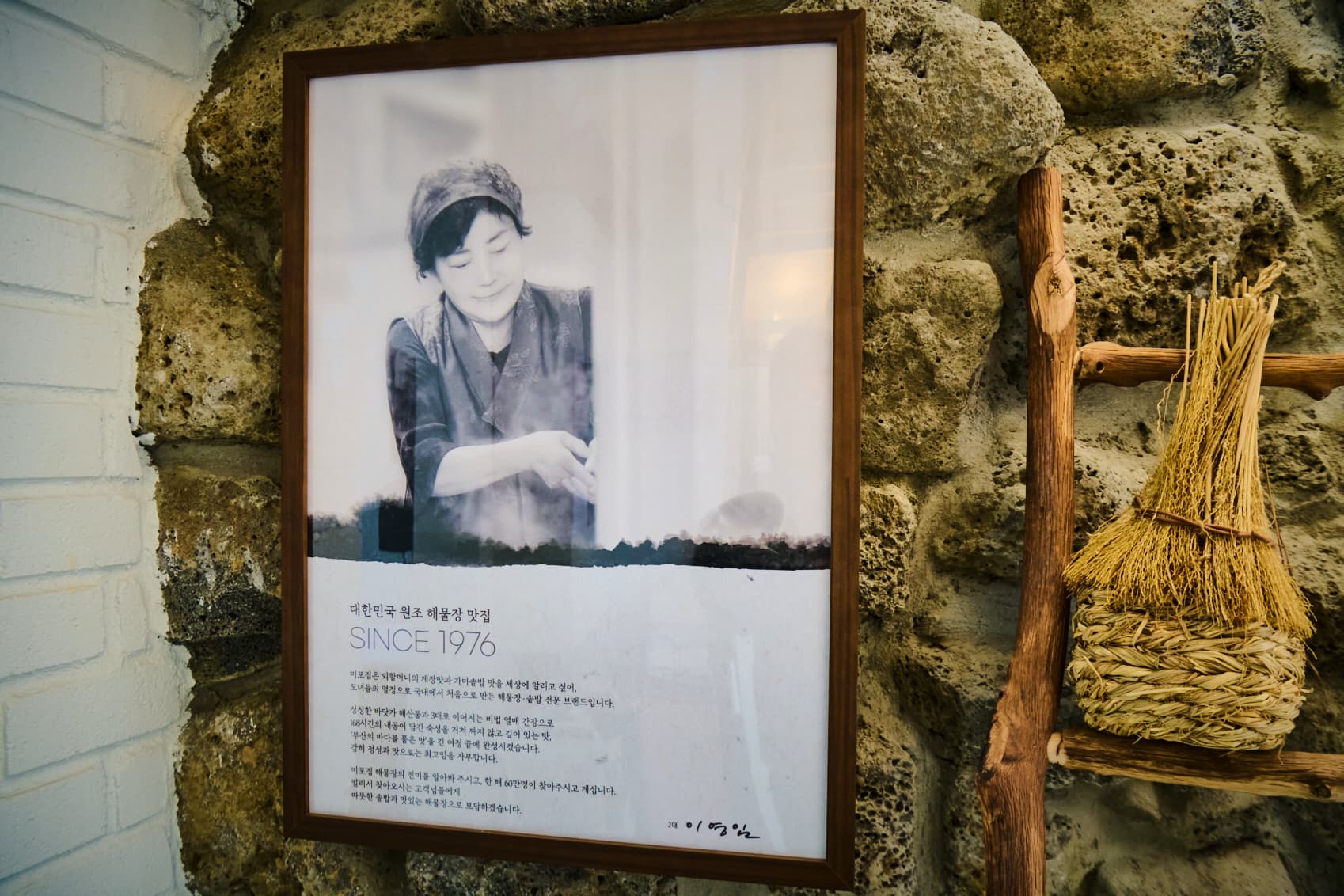 돌담에 걸린 흑백 인물 사진 액자와 짚으로 엮은 도구